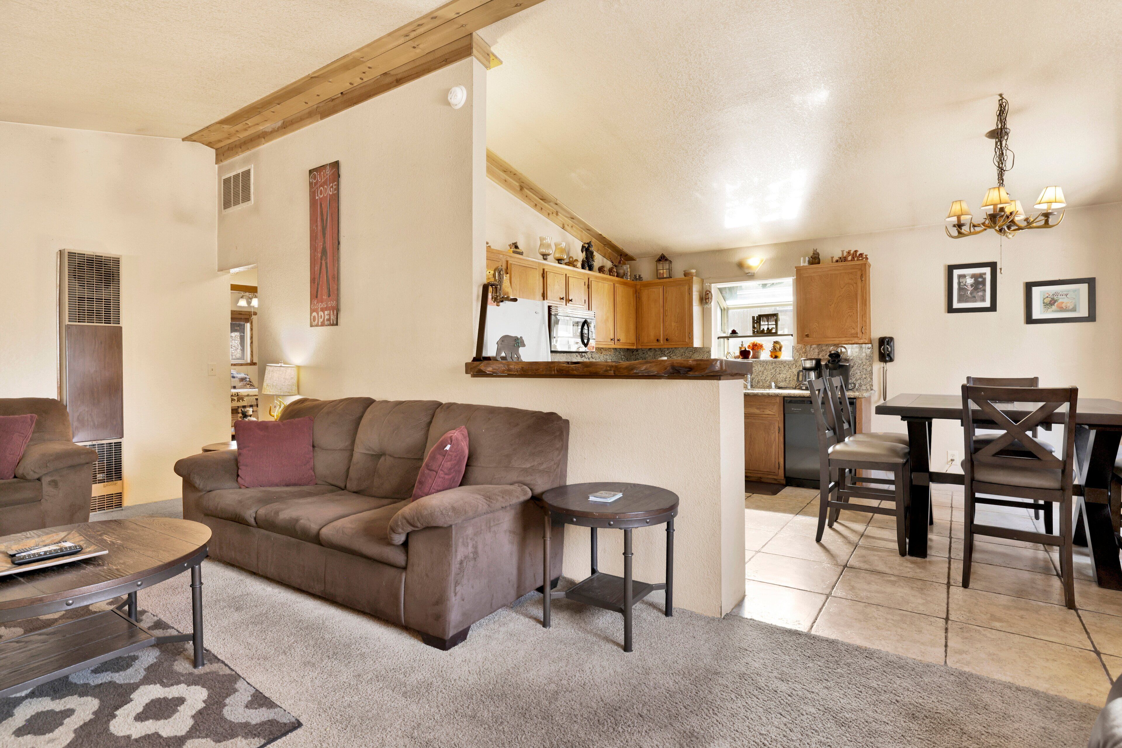 A cozy living room with a brown sofa, a coffee table, and a small side table. A kitchen area with wooden cabinets and a dining table with chairs is visible in the background.