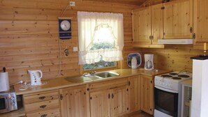 A wooden cabin kitchen with a sink, stove, microwave, kettle, and a clock on the wall.