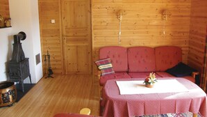 A cozy cabin interior with a wooden wall, a red sofa, a table with a red tablecloth, and a vintage stove.
