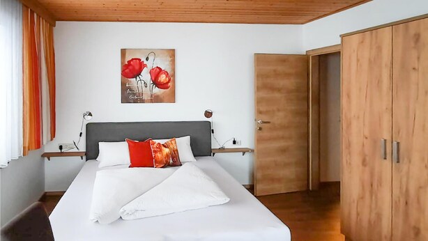 Un lit bien fait, avec du linge de lit blanc et un coussin rouge, une tête de lit en bois et un tableau représentant des fleurs rouges accroché au mur.