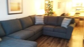 A living room with a dark-colored sofa, two patterned throw pillows, a wooden floor, and a Christmas tree in the background. - Fantastic  Spacious Family Condo (Kelowna)