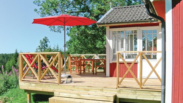 Una terraza de madera con mobiliario exterior, un paraguas rojo y un pequeño edificio con ventanas.