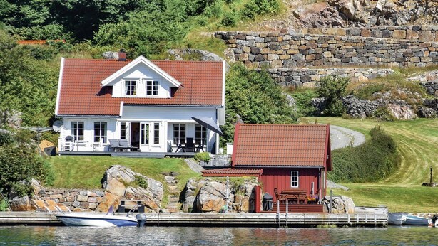 Une maison avec un toit rouge et des murs blancs, un quai avec des bateaux, et un mur de pierre en arrière-plan.
