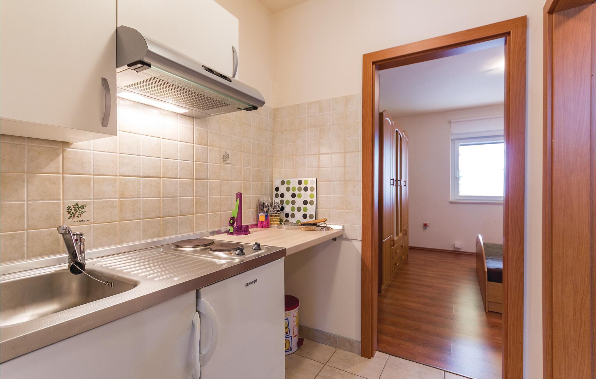 A modern kitchen with a built-in stove, sink, and overhead ventilation. A wooden door leads to a dining area with a window.