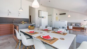 A modern kitchen with a central island, white dining table with red placemats, and a living area with a sofa and striped cushions.