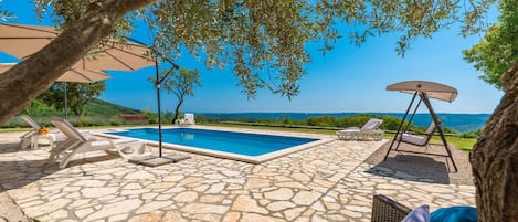 Un espace piscine doté de chaises longues et d’une chaise suspendue, entouré d’un sol en pierre et d’arbres.