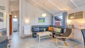 Un salon chaleureux avec un canapé, des fauteuils et une table basse. Un tableau orne le mur, un luminaire est suspendu au plafond et une fenêtre est agrémentée de rideaux.