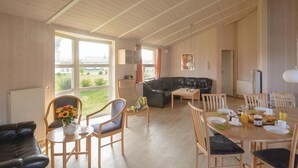 A spacious living room with a black leather sofa, wooden dining table, and chairs. A large window offers a view of the outdoors. - Freibeuterweg 6 - Dorf 6 (Travemünde-Priwall)