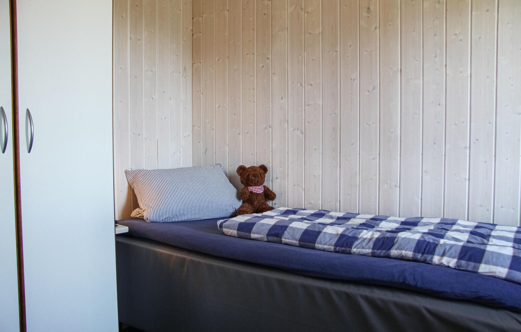 A small, single-bed room with a wooden wall, a striped pillow, a checkered blanket, and a teddy bear.