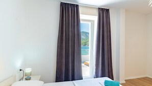 A hotel room with a bed, a nightstand, a lamp, and a view of the sea through a window with curtains.