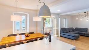A modern living room with a wooden dining table, a grey sofa, and a large window offering a view of the outdoors.