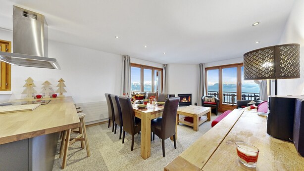A modern dining area with a wooden table, chairs, and a fireplace. There are windows with a view of mountains.