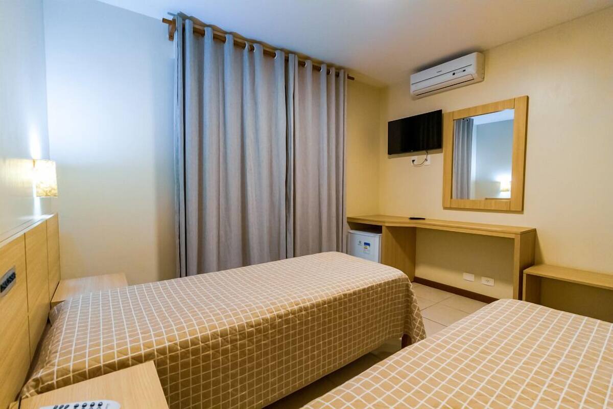 A hotel room with two beds, a desk, a mirror, an air conditioning unit, and a lamp.