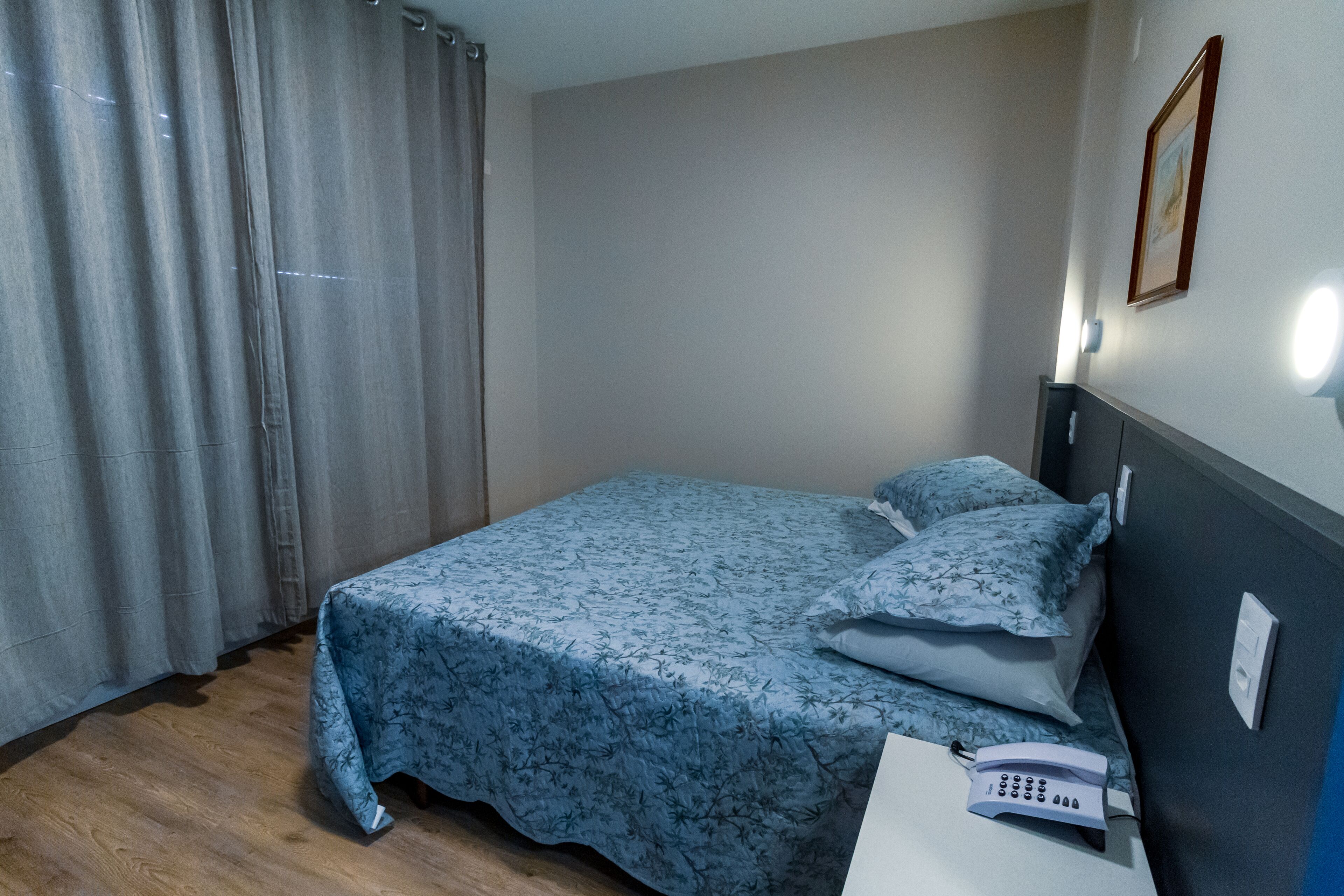 Un lit simple avec une couvre-lit à motifs floraux, un téléphone sur la table de chevet et un luminaire fixé au mur.