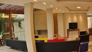 A modern living room with a flat-screen TV, comfortable seating, and a glass wall offering a view of palm trees. - A Furninha Hotel (Torres)