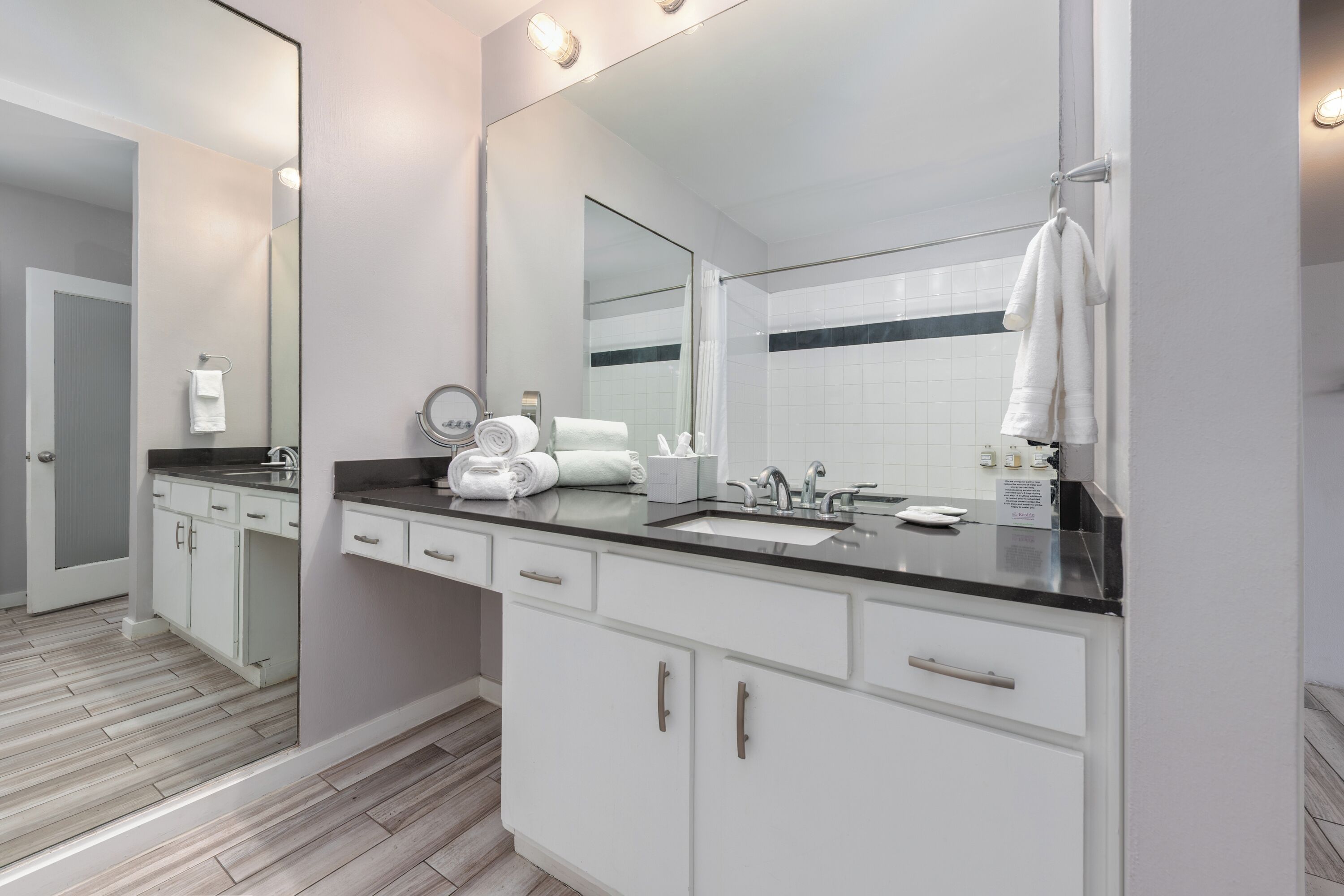 A modern bathroom with a large mirror, white cabinets, a black countertop, and a shower area.