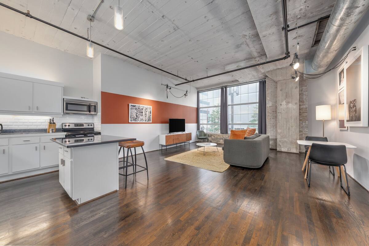A modern kitchen with a central island, a dining area with a gray sofa, and a large window providing natural light.