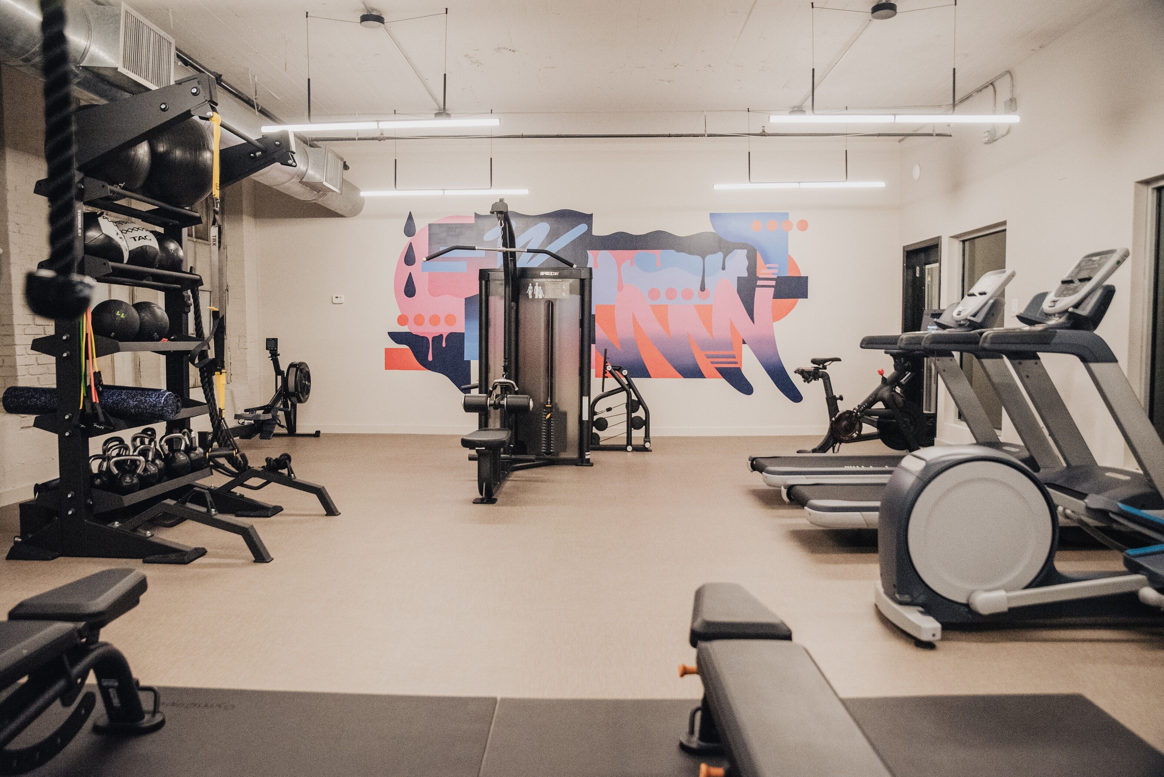 Een fitnessruimte met trainingsapparatuur, een loopband en een muur met abstracte kunst.