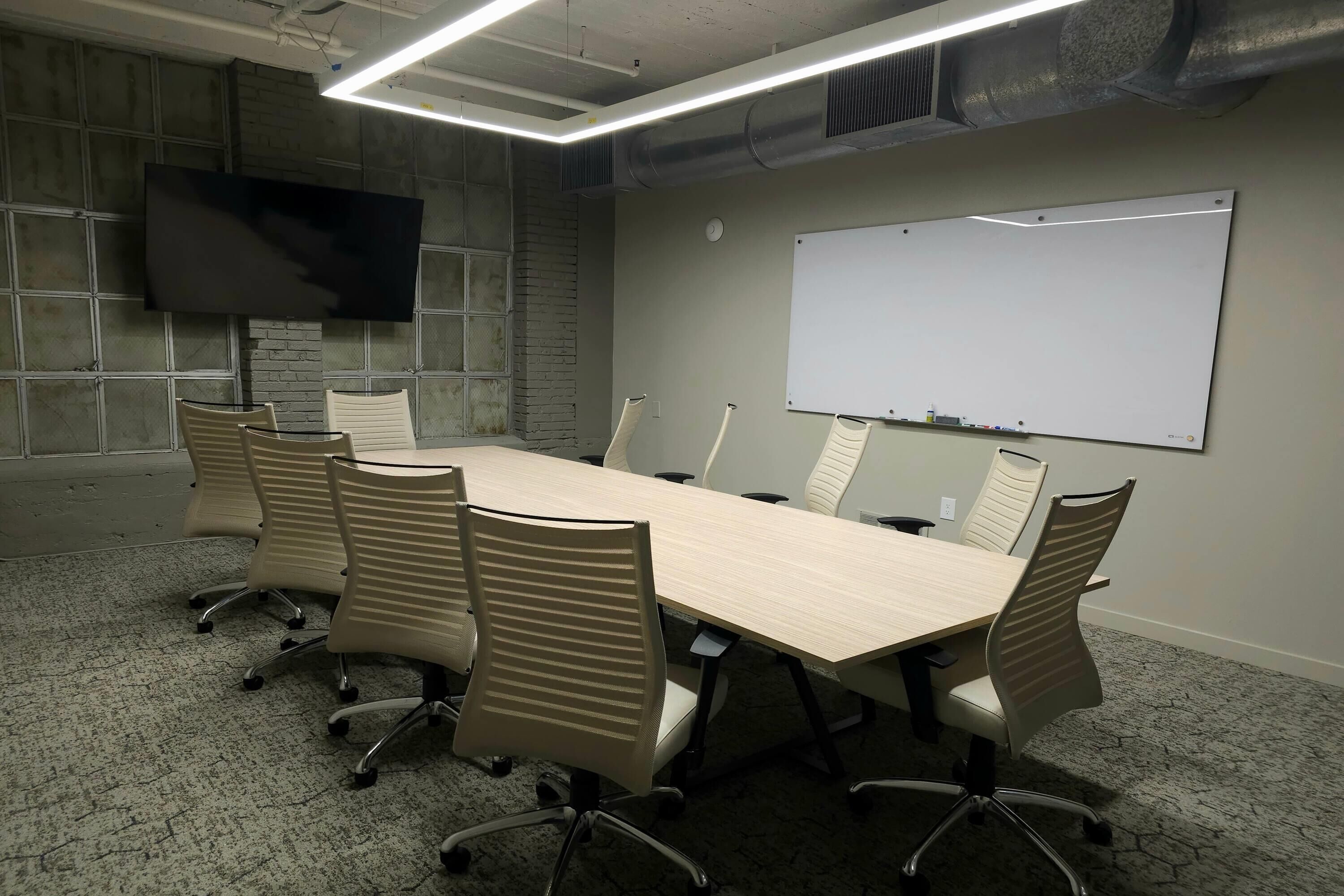 A conference room with a long table, six chairs, a whiteboard, and a flat-screen TV mounted on the wall.