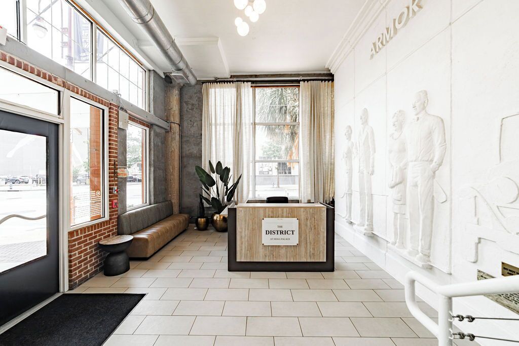 A reception area with a wooden reception desk labeled 'The District,' large windows, a brick wall, and a wall-mounted sculpture.