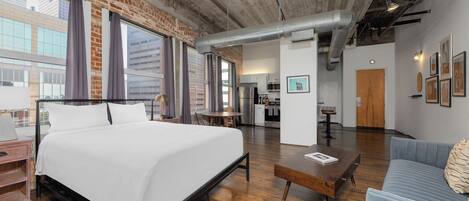 Une chambre d’hôtel moderne avec un grand lit, un canapé, une table basse en bois et, au fond, une petite cuisine.