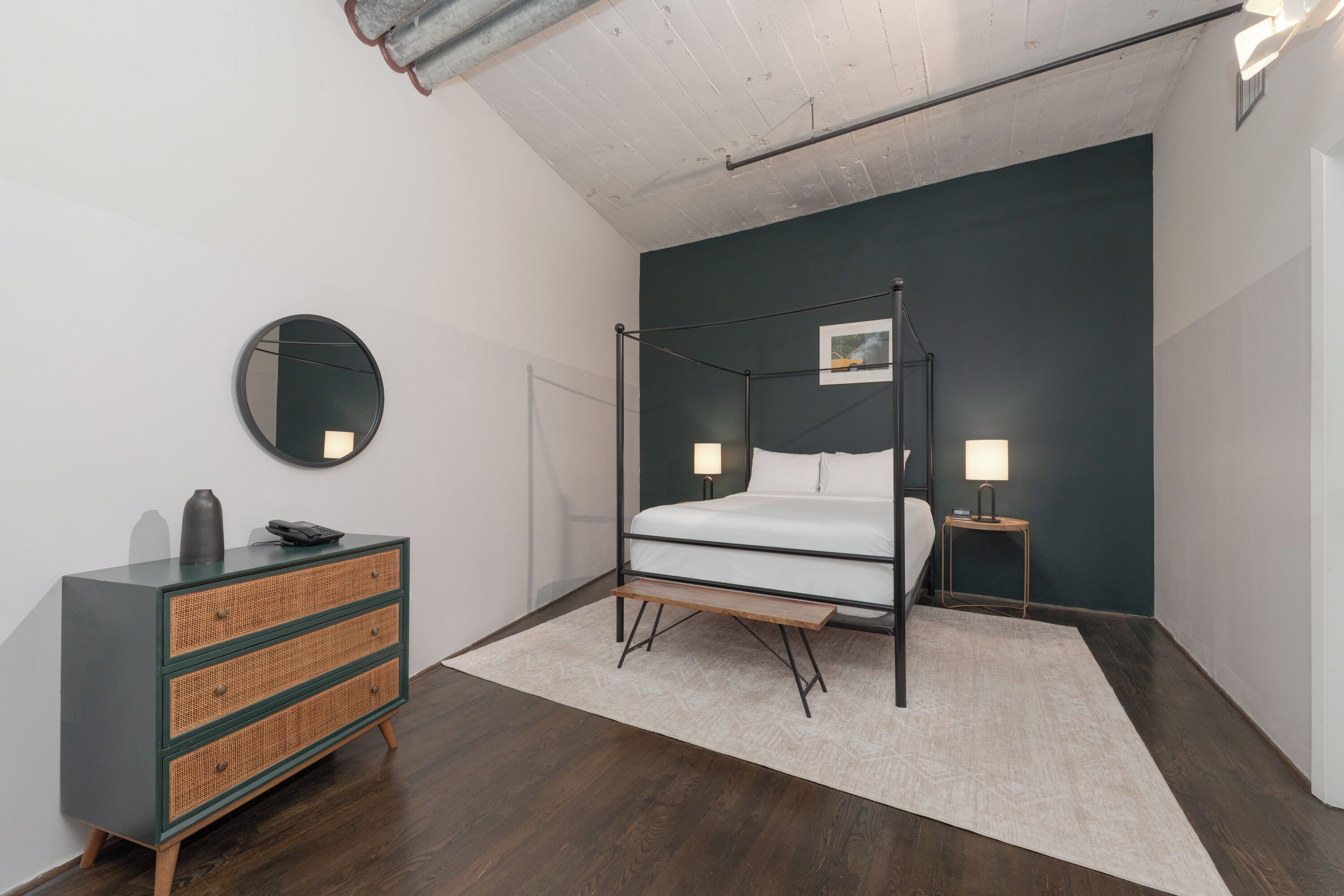 Ein modernes Schlafzimmer mit einem Himmelbett, einem Nachttisch mit Lampe, einer Kommode mit Spiegel und einem kleinen Bilderrahmen an der Wand.