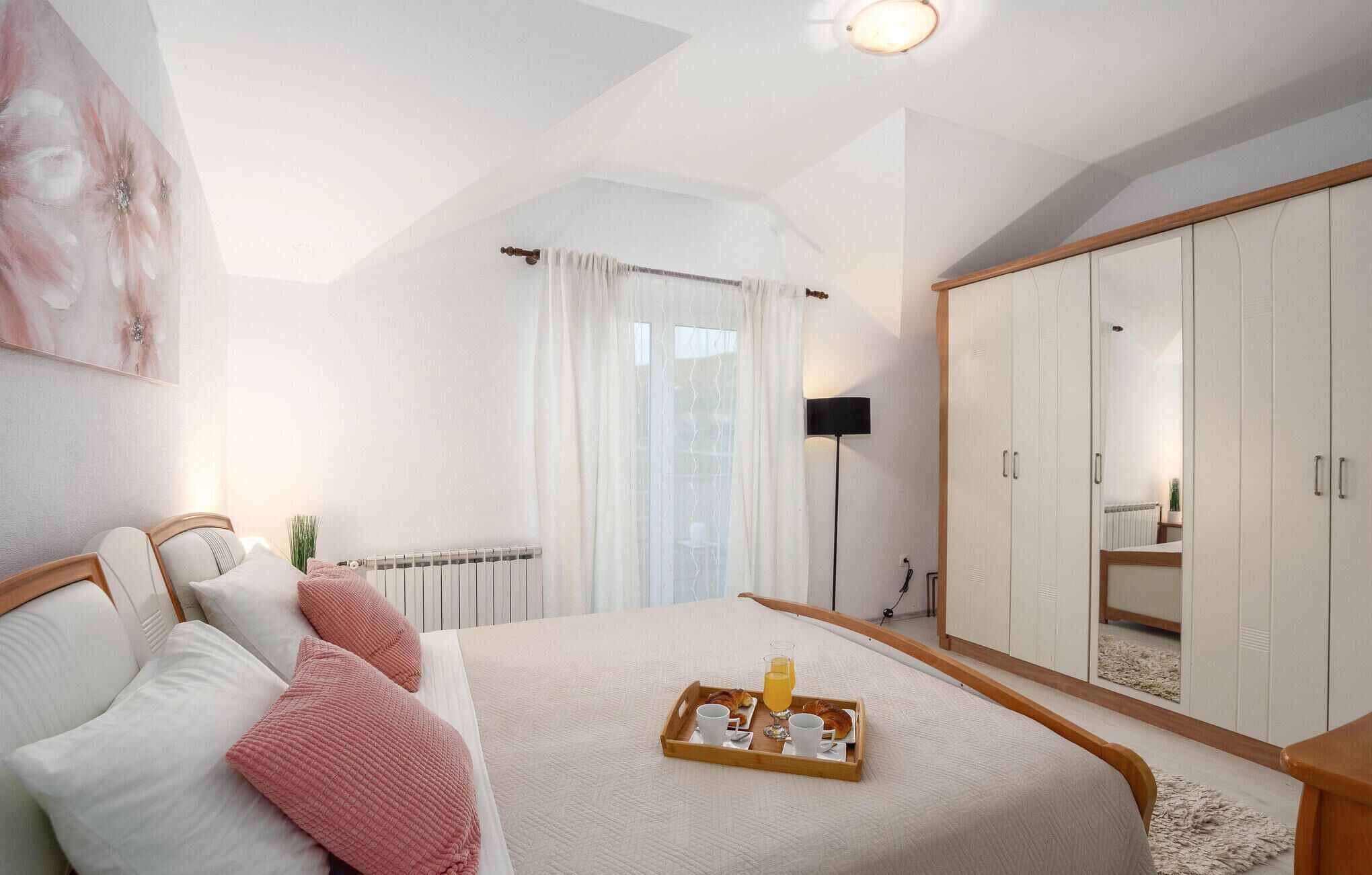 Een slaapkamer met een bed, een nachtkastje met een ontbijtje erop, een kledingkast, een radiator en een raam met gordijnen.