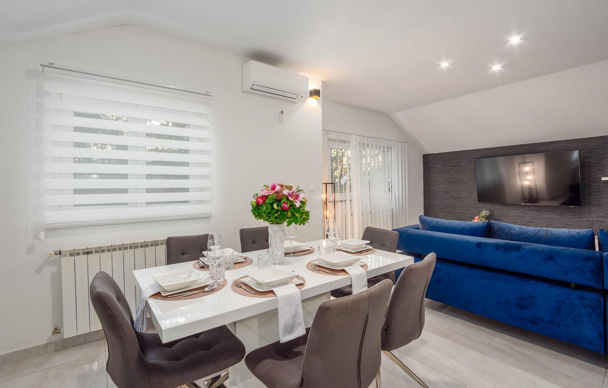 Un comedor moderno con una mesa blanca dispuesta para cuatro, un sofá azul y un televisor de pantalla plana montado en la pared.