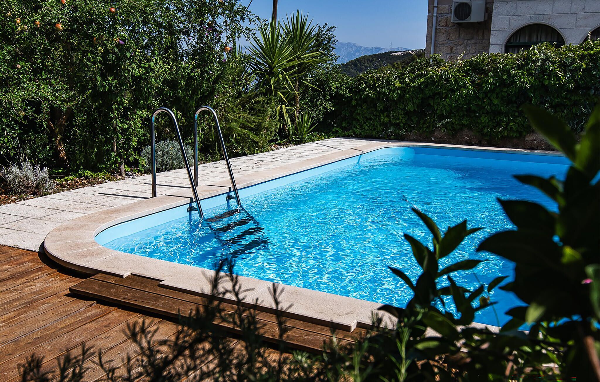 Una piscina rectangular amb un solàrium de fusta i escales metàl·liques, envoltada de vegetació abundant i un edifici de pedra al fons.