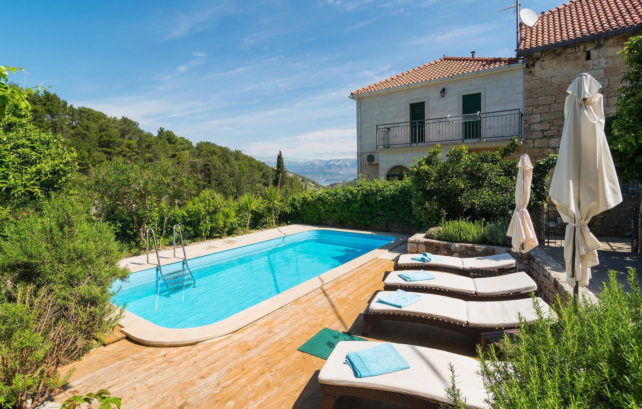 A swimming pool with lounge chairs and a wooden deck, surrounded by greenery and a stone building with a balcony.