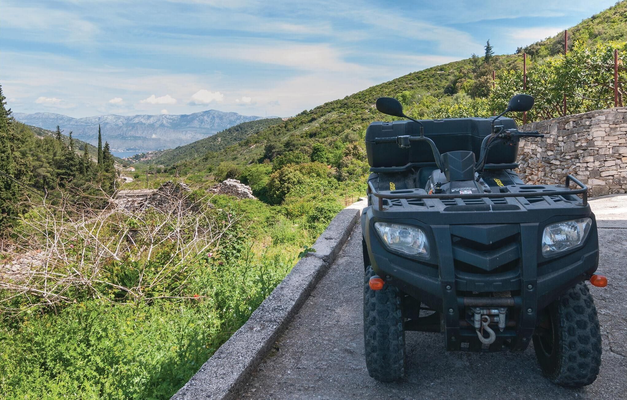 A quad bike parked on a paved path with a stone wall on one side and a mountainous landscape in the background.
