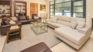 A spacious living room with a brown leather sofa, a beige sofa, and a glass coffee table. There are bookshelves, a painting, and a large window with a view of trees.