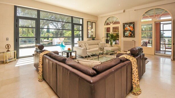 A spacious living room with a large sectional sofa, a glass coffee table, and a beige sofa. There are large windows offering a view of the outdoors.