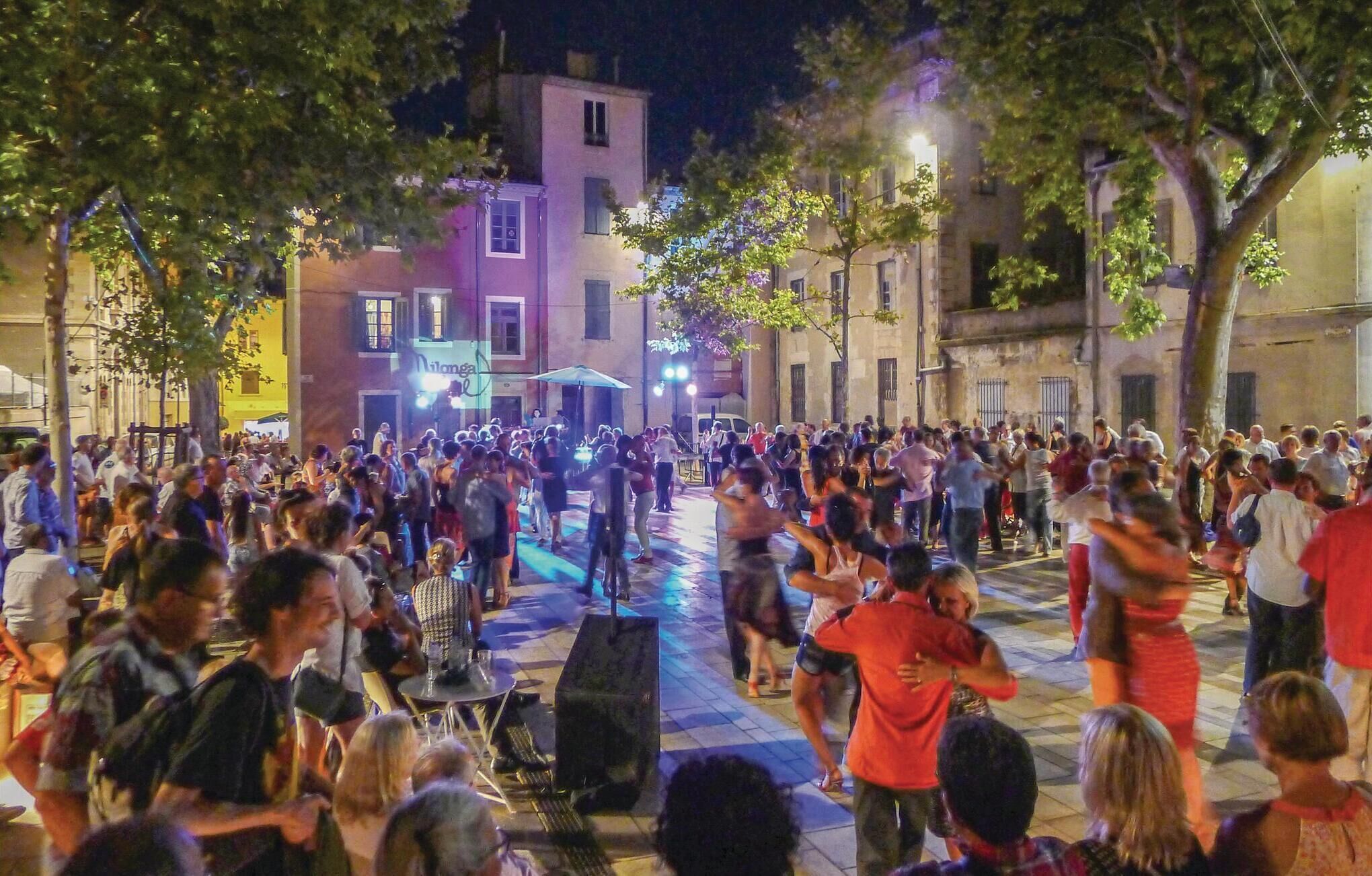 A crowded outdoor event with people gathered around a central stage, surrounded by buildings.