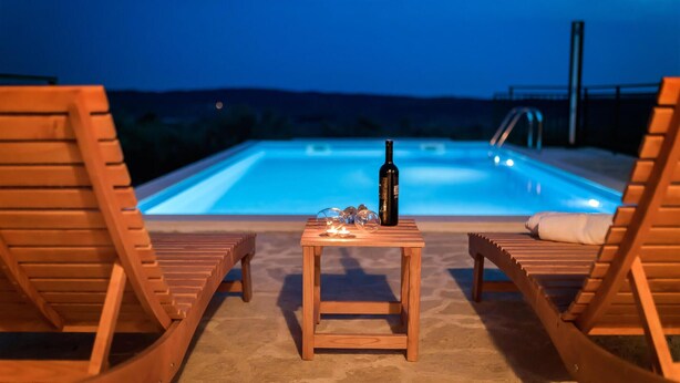 Ein Poolbereich mit Holzliegen, einem kleinen Tisch mit Flasche und Gläsern und Blick auf ein Schwimmbad bei Nacht.