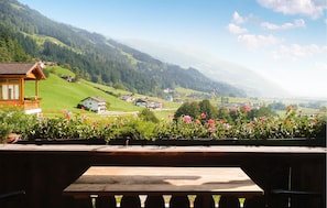 Una mesa y sillas de madera en un balcón con vista a un pueblo, casas y colinas verdes al fondo.