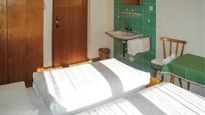 Ein kleines, sauberes Zimmer mit einem Einzelbett, einem Holzstuhl, einem grünen Tisch und einem Badezimmer mit Waschbecken und Spiegel.