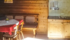 Ein Holzblockhaus-Interieur mit einem Esstisch, Stühlen und einer Bank. Es gibt eine Küchenzeile mit darüber hängenden Küchenutensilien.