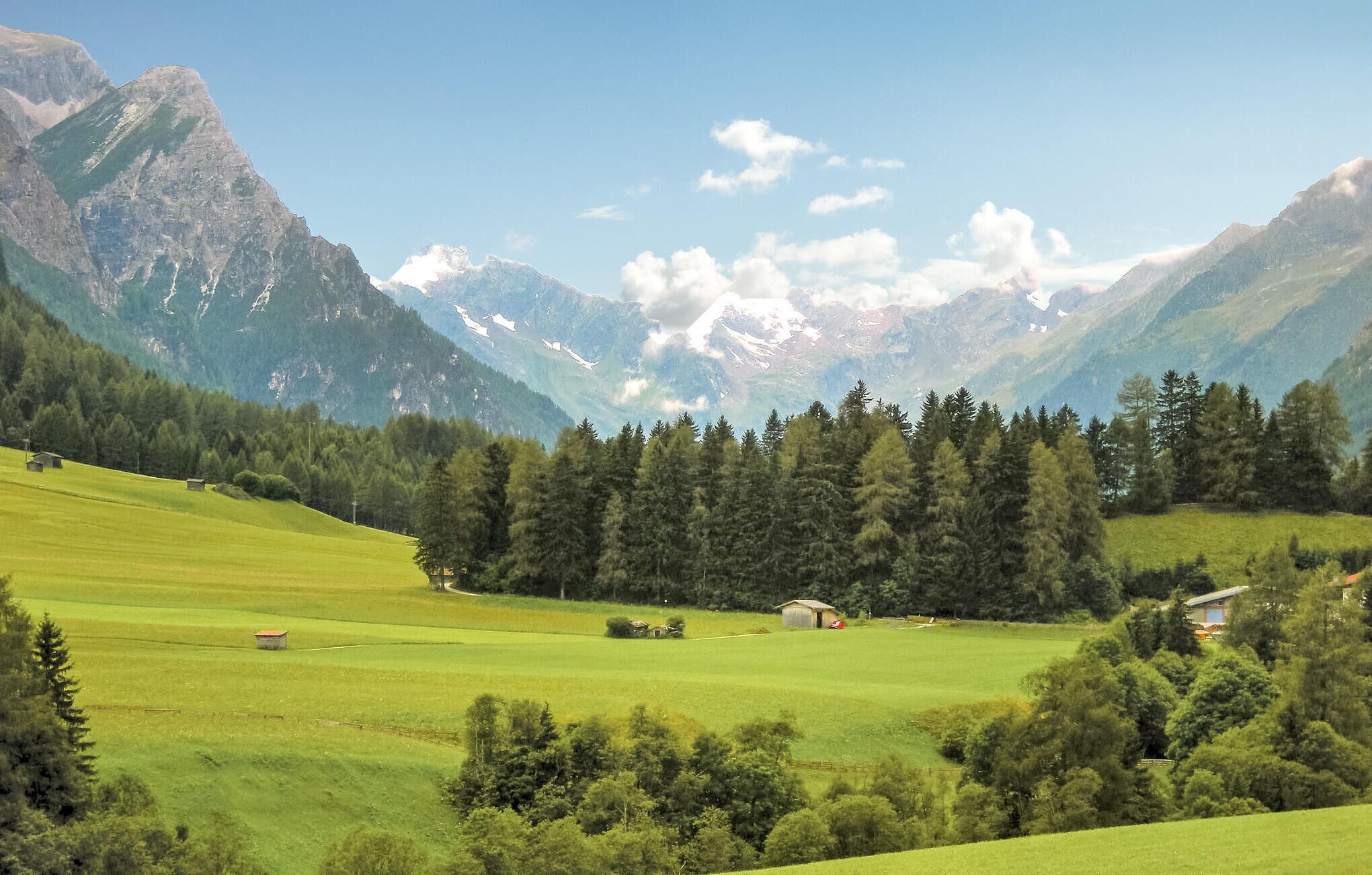 A mountainous landscape with green fields, trees, and a few small buildings.