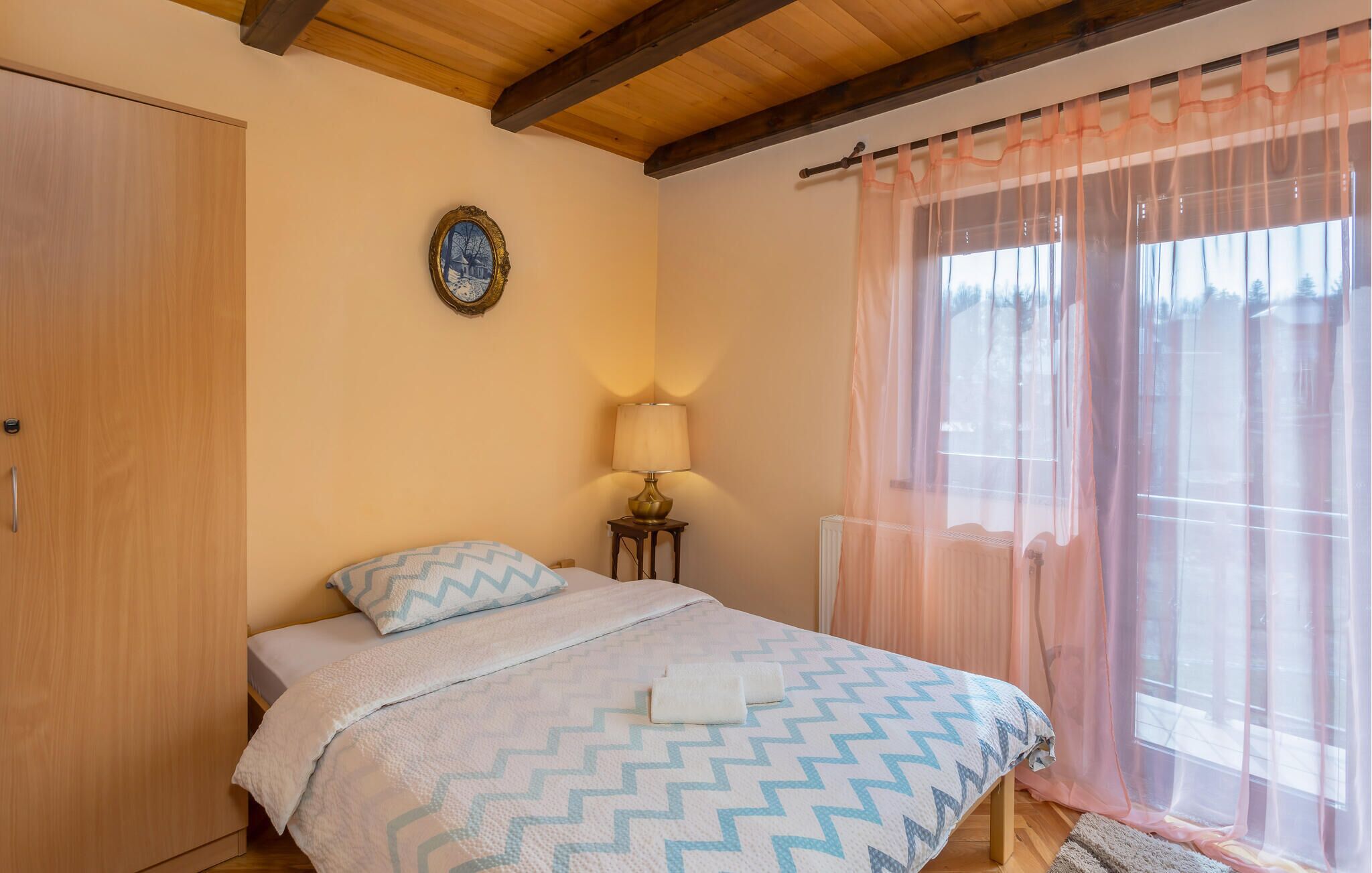 Une chambre à coucher comprenant un lit, une table de chevet, une lampe, une fenêtre avec des rideaux légers et un plafond en bois.