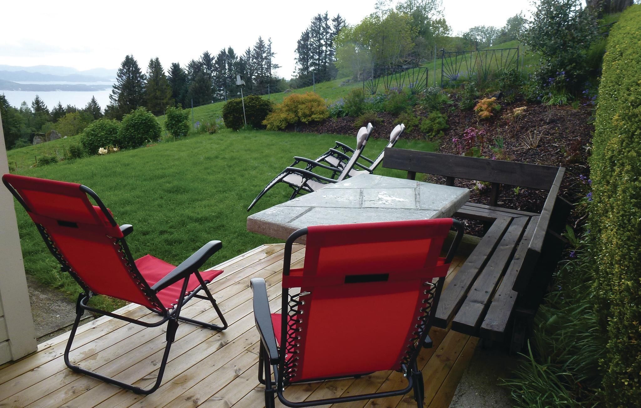 A wooden deck with a table and red chairs, overlooking a well-maintained garden and a view of the water.