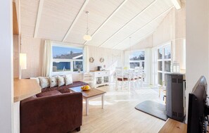A spacious living area with a brown sofa, a dining table, and a TV. There are large windows and a view of the outside.