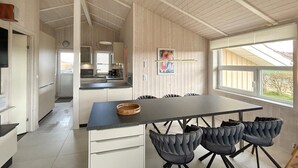 A modern kitchen with white cabinets, a dining area with a large table and upholstered chairs, and a view through a window.
