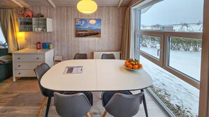 Spiseplads med et hvidt bord, fire stole, en skål med frugt, et billede og udsigt til et sneklædt landskab gennem store vinduer.