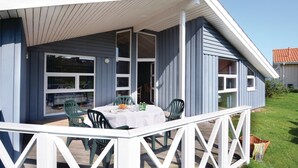Un espacio exterior techado con una mesa y sillas, una baranda blanca, y vista del interior de la casa a través de grandes ventanales.