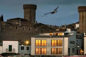 A historic building with a tower, modern buildings with illuminated windows, and a body of water with boats. - AMAZING APARTMENT SEA VIEWS IN TOSSA (Tossa de Mar)