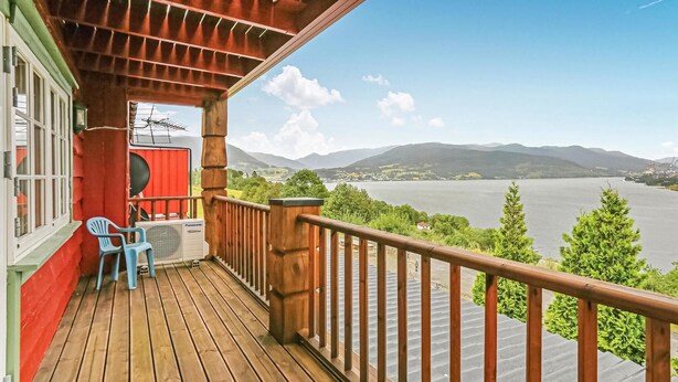 A wooden deck with a red railing, a blue chair, and a red speaker on a balcony overlooking a lake and mountains. - 10 bedroom amazing home in ølen (Ølen)