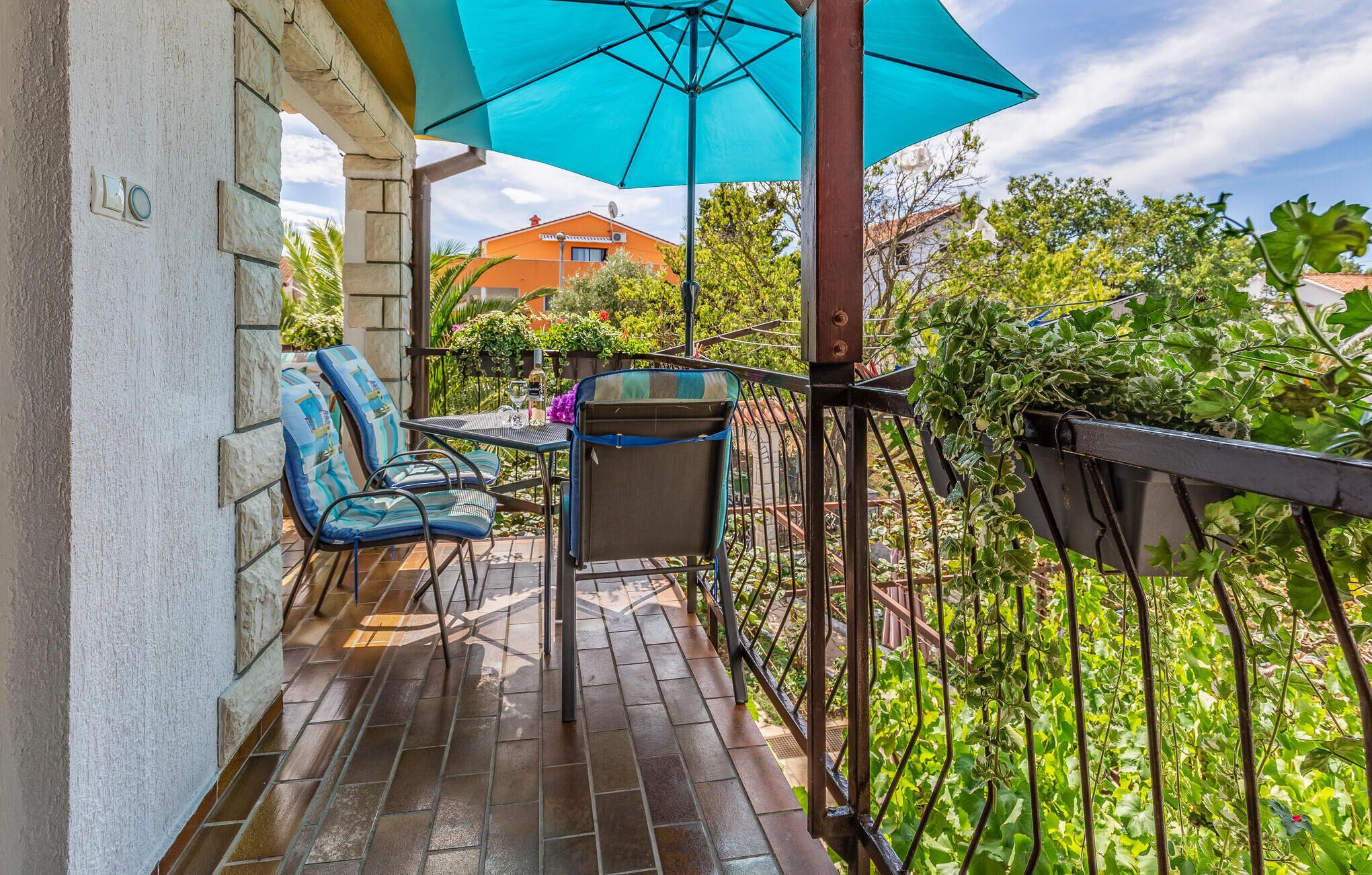 Un balcon avec deux chaises et une table, un parasol bleu, et une vue sur un jardin et des bâtiments.
