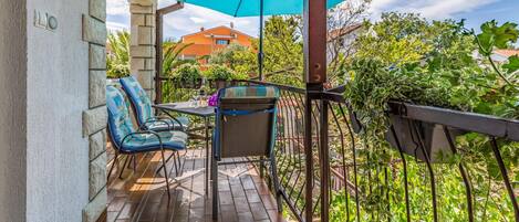 Un balcon avec deux chaises et une table, un parasol bleu, et une vue sur un jardin et des bâtiments.