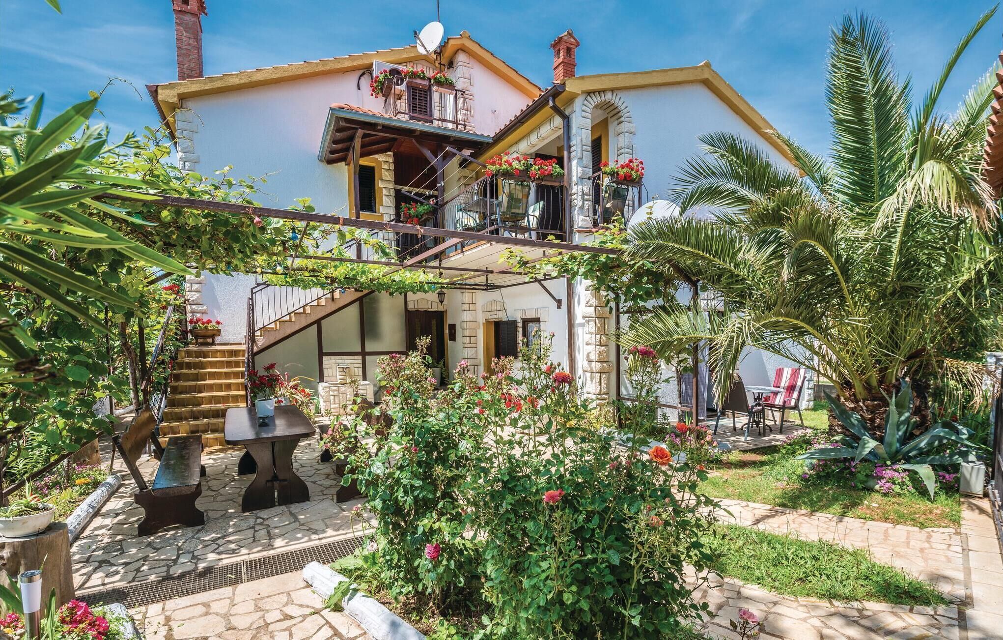 A two-story house with a balcony, a stone staircase, and a garden with various plants and flowers.